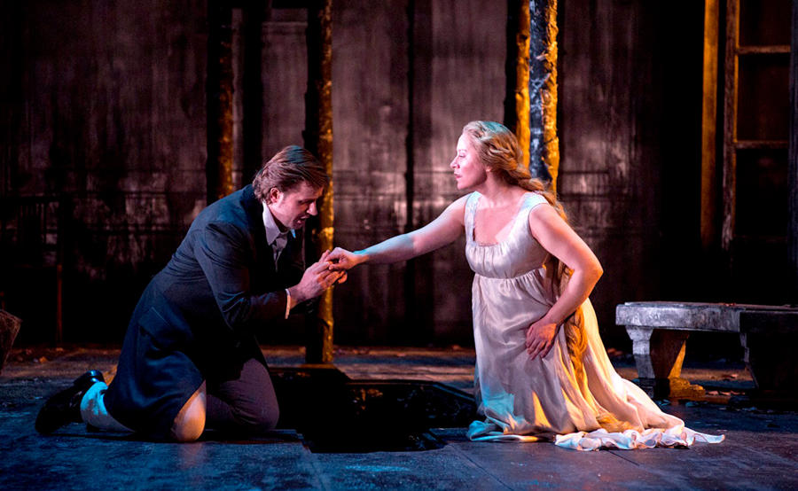 A white man and a white, blonde woman are on their knees on stage. He holds her right hand with both his hands, looking at it, while she looks at him. 
