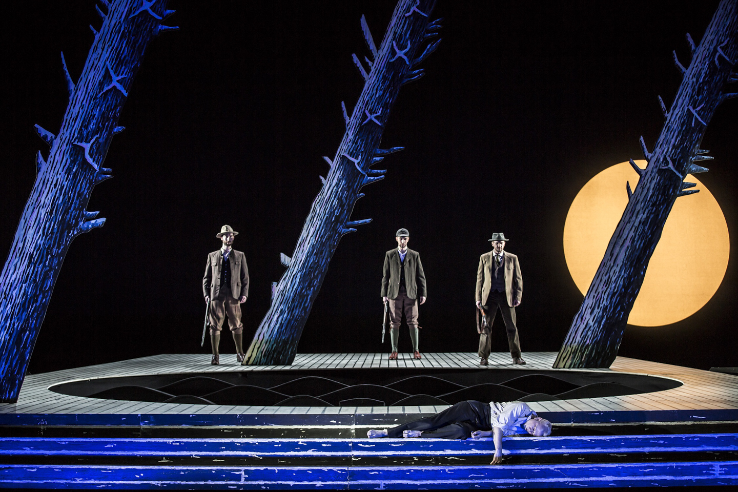 Three men in hunting suits among the trees, looking down at a man lying on the ground in Rusalka