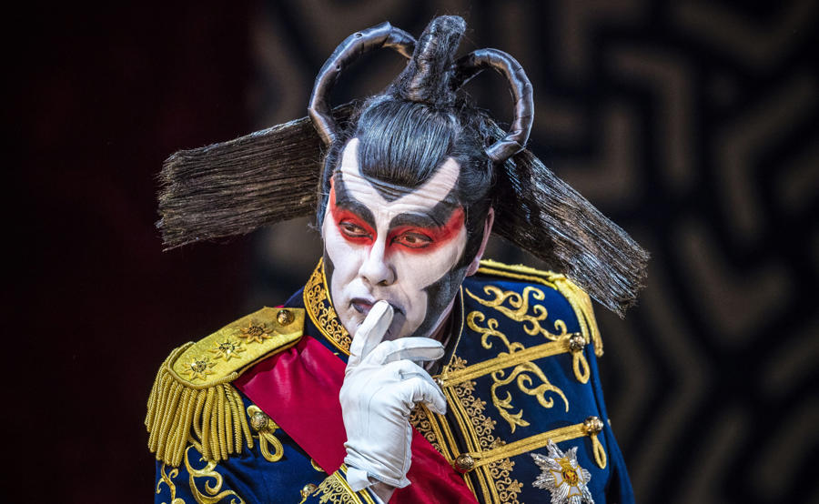 Stephen Richardson as The Mikado, wearing an elaborate black wig and blue and gold military jacket, with his hand to his mouth