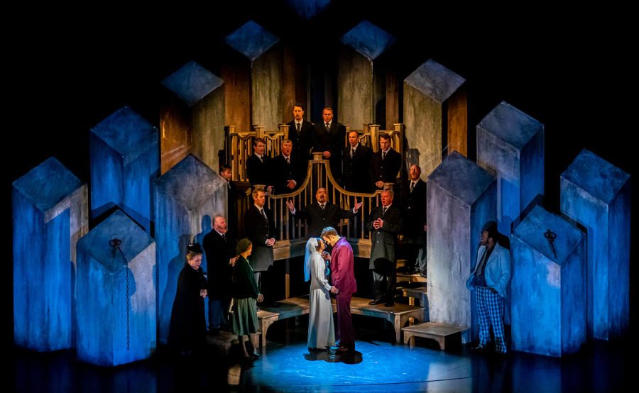 A choir sings on stage behind a white woman and a white man holding hands under the spotlight. 