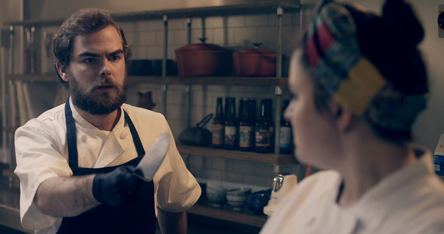 Angus (Arthur Bruce) pointing a sharp chef's knife at Belle (Charlie Drummond) in The Narcissistic Fish