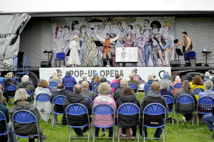 Audience from behind watching Pop-up Opera at Wigtown Book Festival, seated on blue plastic chairs, with the performers on the back of a lorry decorated with an illustration of characters from the show