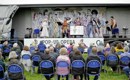 Audience from behind watching Pop-up Opera at Wigtown Book Festival, seated on blue plastic chairs, with the performers on the back of a lorry decorated with an illustration of characters from the show