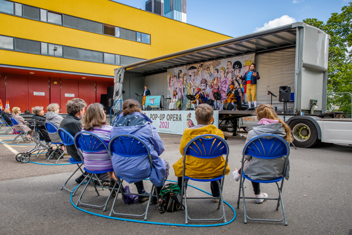 Audience members from behind watching a performance of Pop-up Opera taking place on a lorry, seated on blue plastic chairs and wearing raincoats
