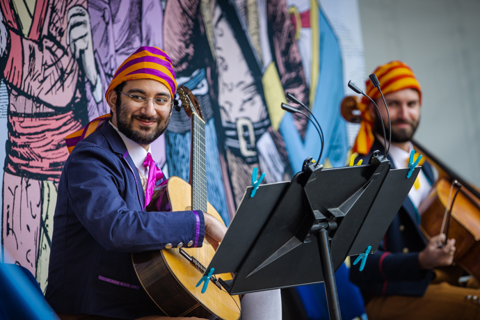 Sasha Savaloni in Pop-up Opera smiles towards the camera, holding an acoustic guitar, wearing a yellow and purple head dress.