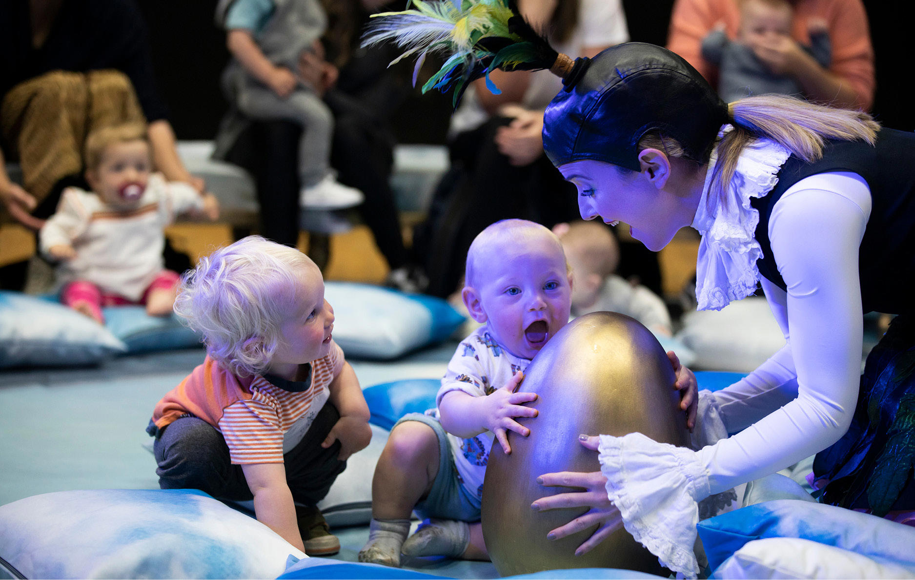 A performance of BambinO. A woman holds a large golden egg, wearing a head dress with plumes of feathers sticking out the top. A small child also holds the egg, while another toddler gazes up at the woman.
