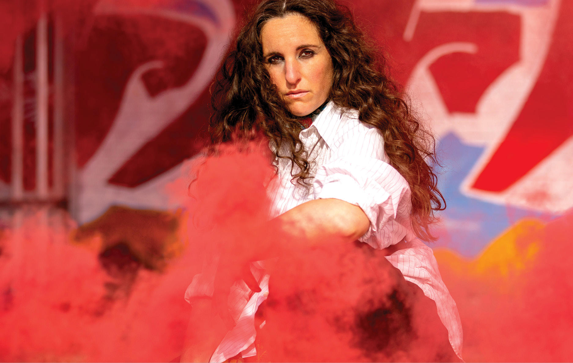 A woman, Carmen, with long, curly hair wearing a white shirt, surrounded by red smoke