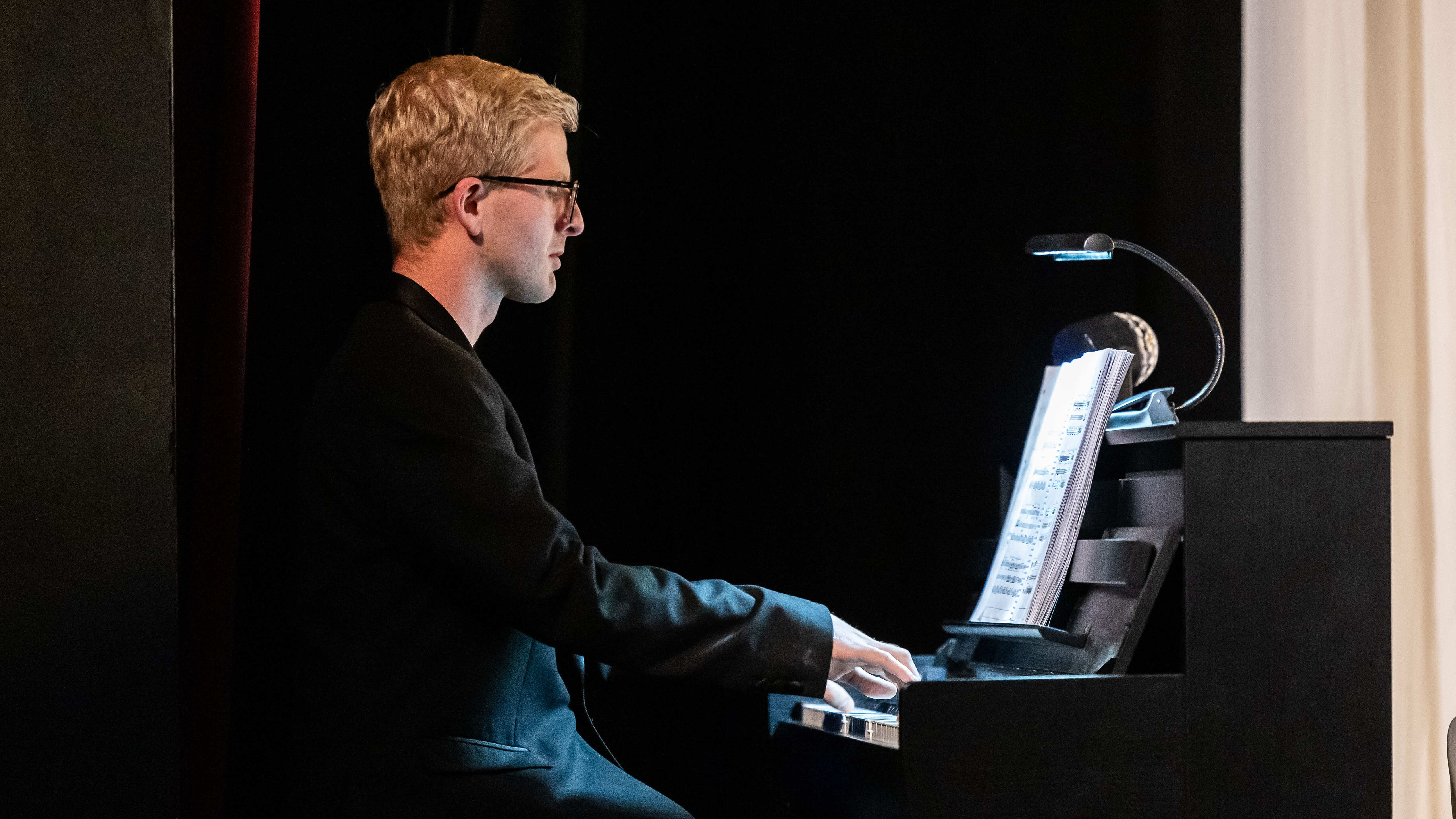 A white, blonde man with glasses plays the piano.