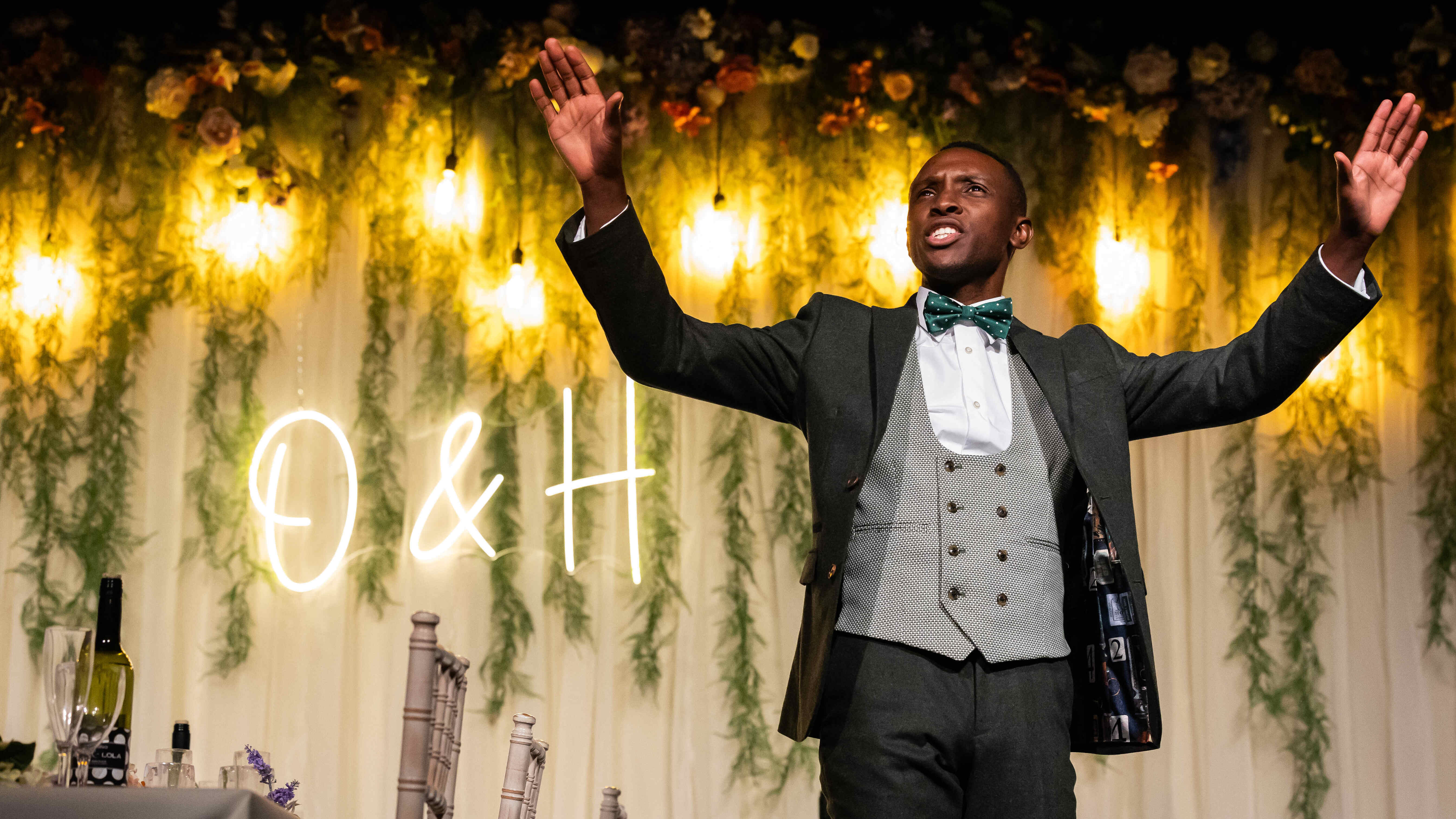 A black man wears a suit and sings with his hands in the air.