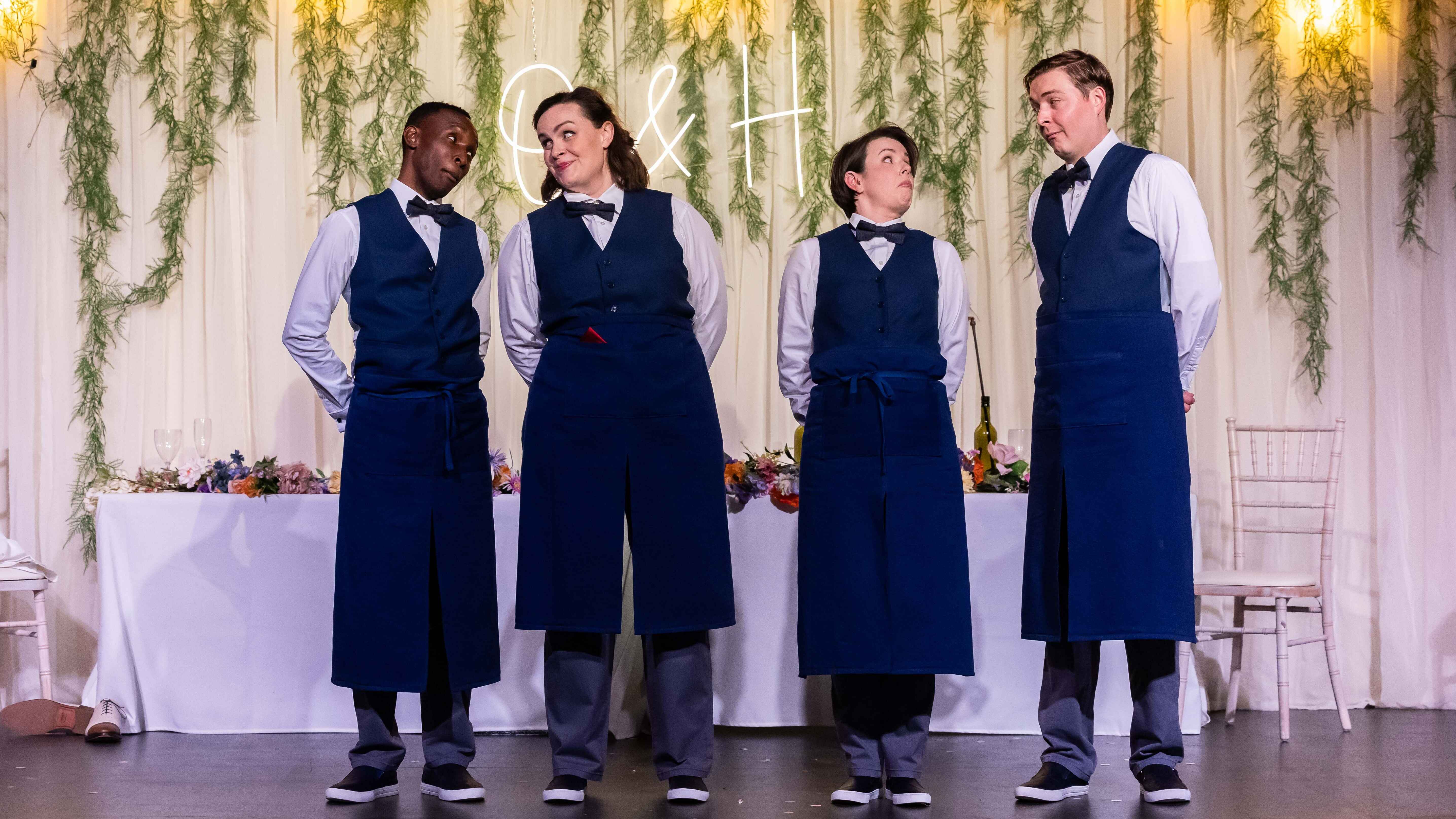 Four cast members dressed as waiters look at one another suspiciously.