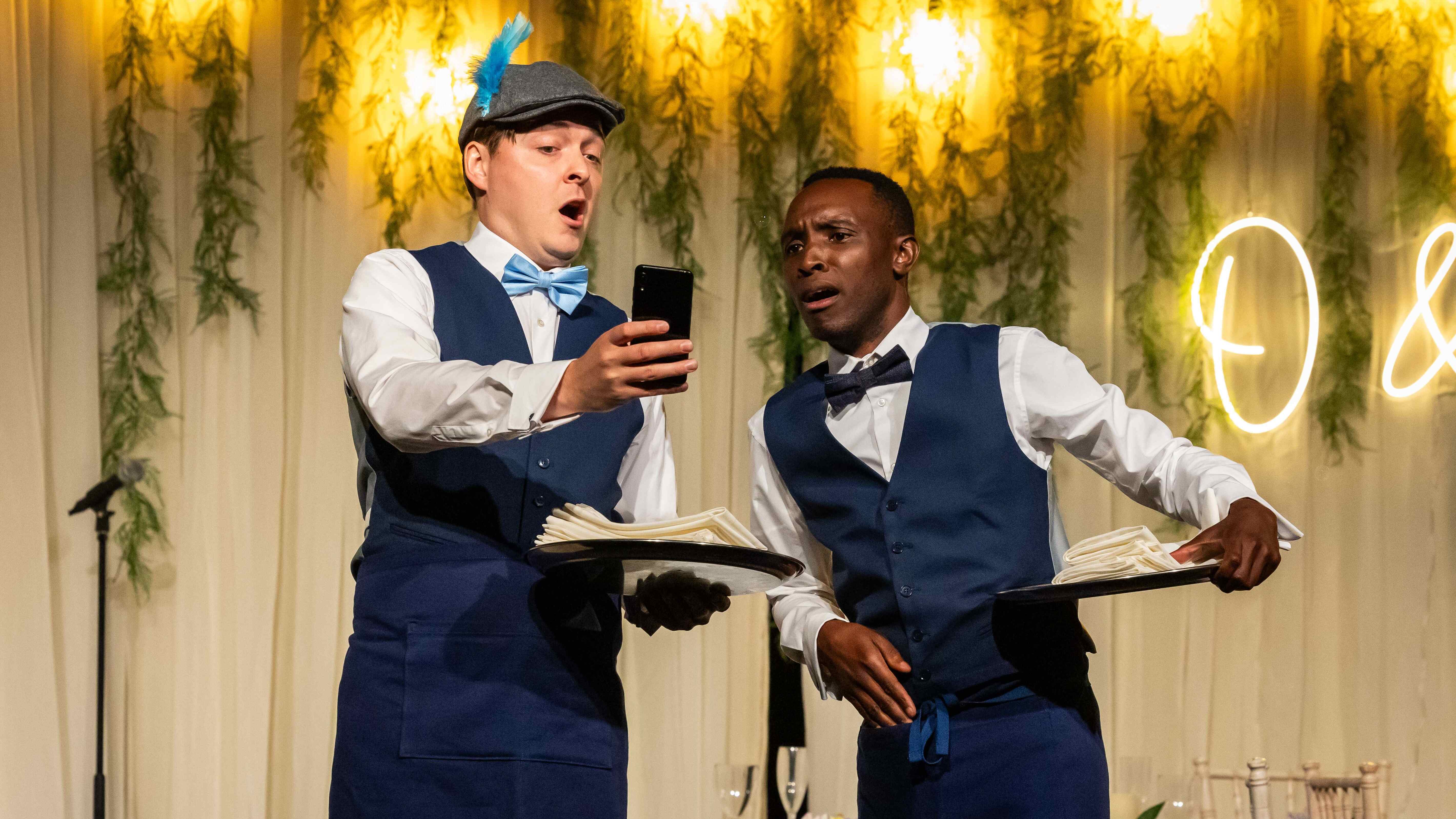 A white man shows his phone to a black man. Both are dressed as waiters.