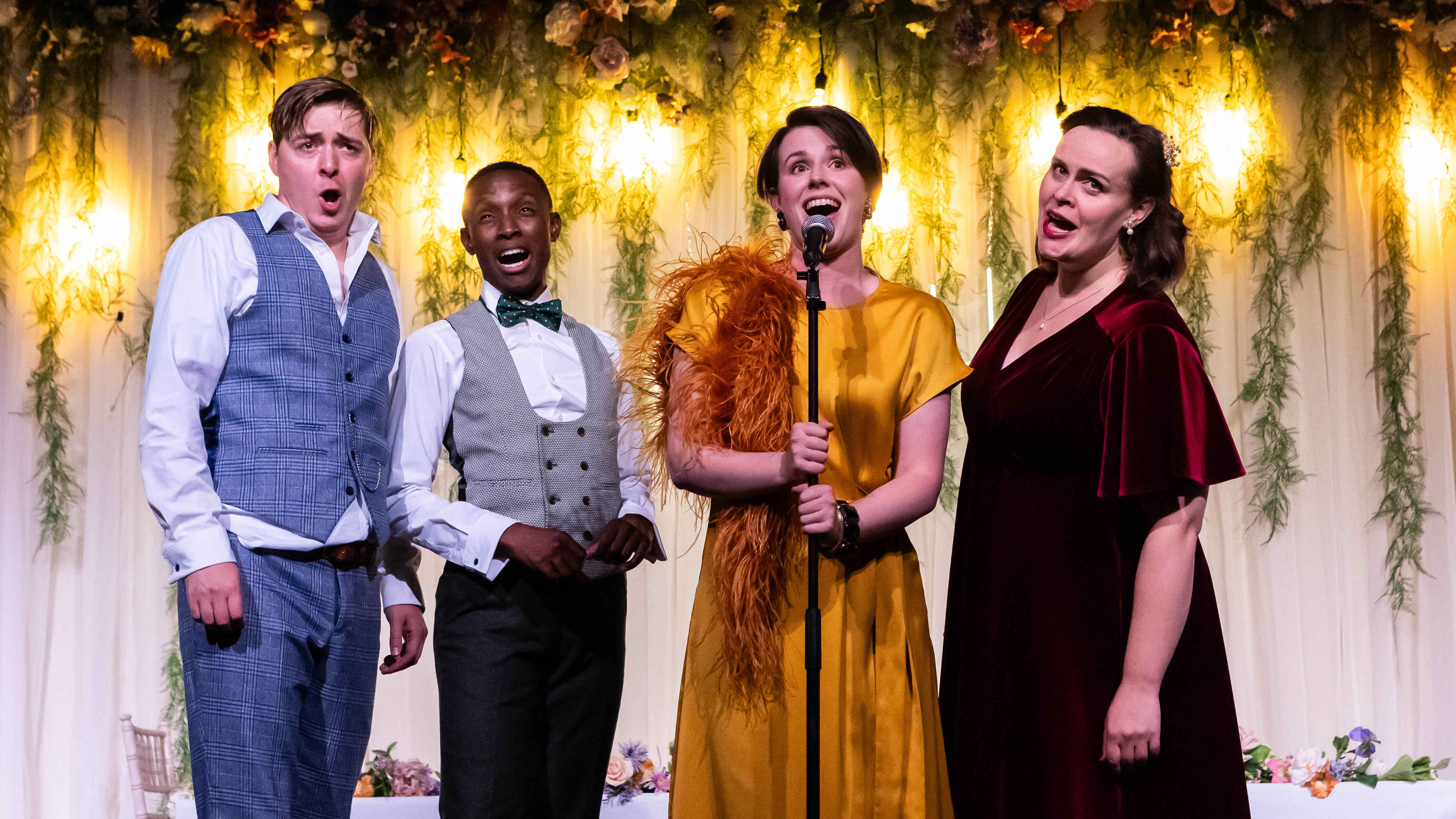 Two men and two women in fancy dresses sing. One holds a microphone.