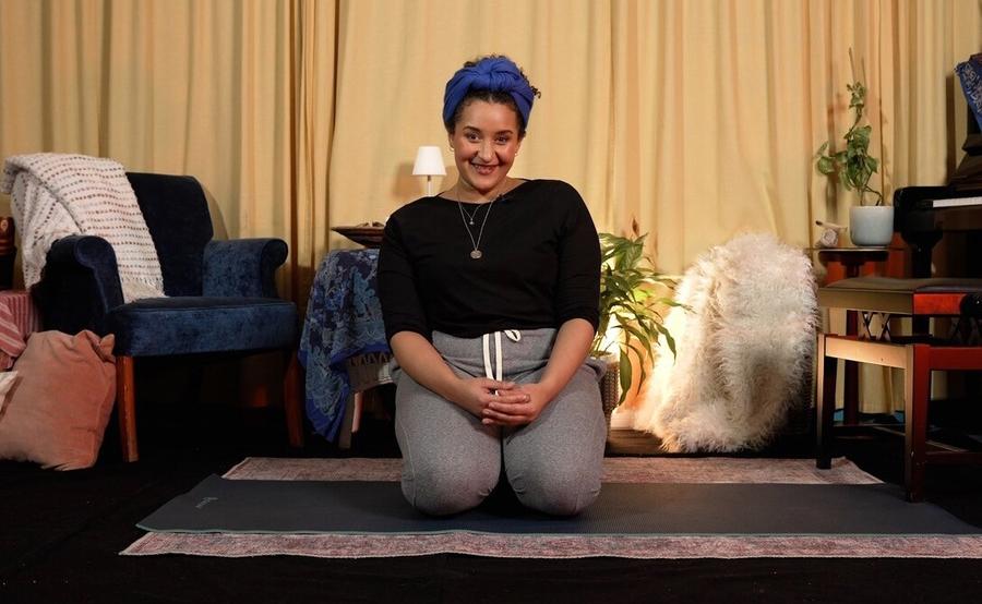 A portrait of Lea Shaw. She is smiling to the camera and kneeling on a yoga matt.